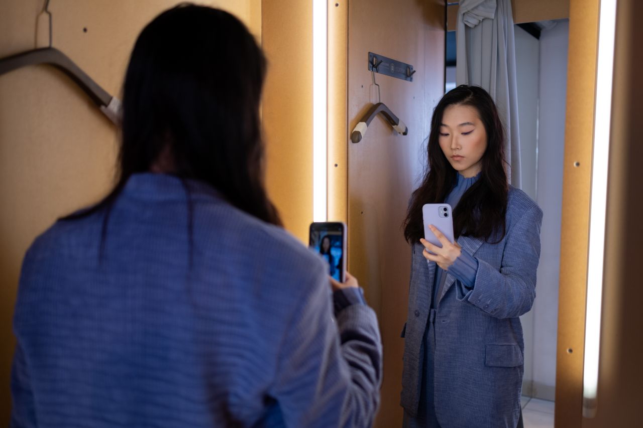 A stylish Asian woman takes a selfie in a fitting room with a smartphone, wearing a fashionable blazer.